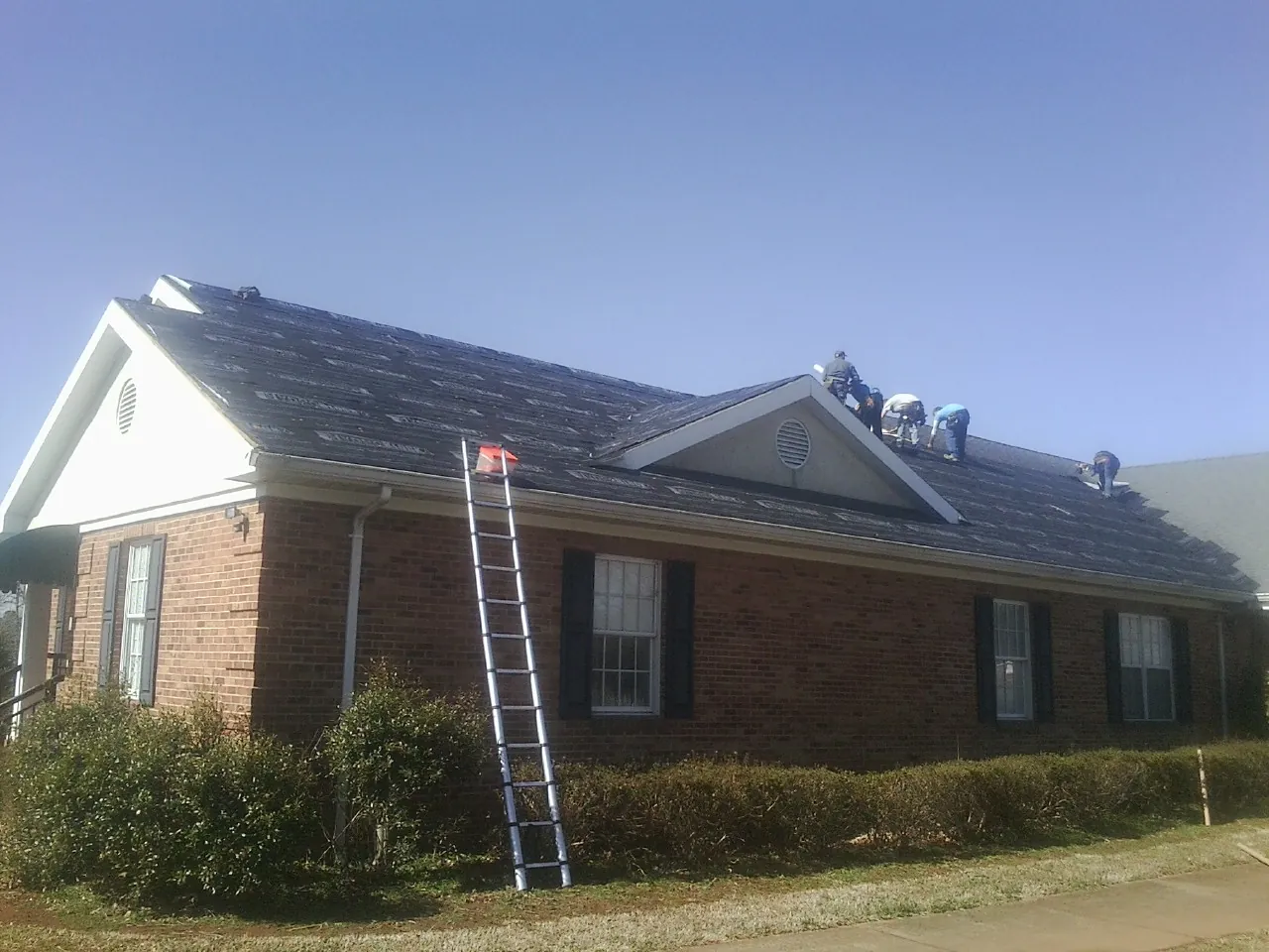Crew tearing shingles off a brick ranch roof with a ladder set — Metal Roof Repair work by our crew in Wells Bridge