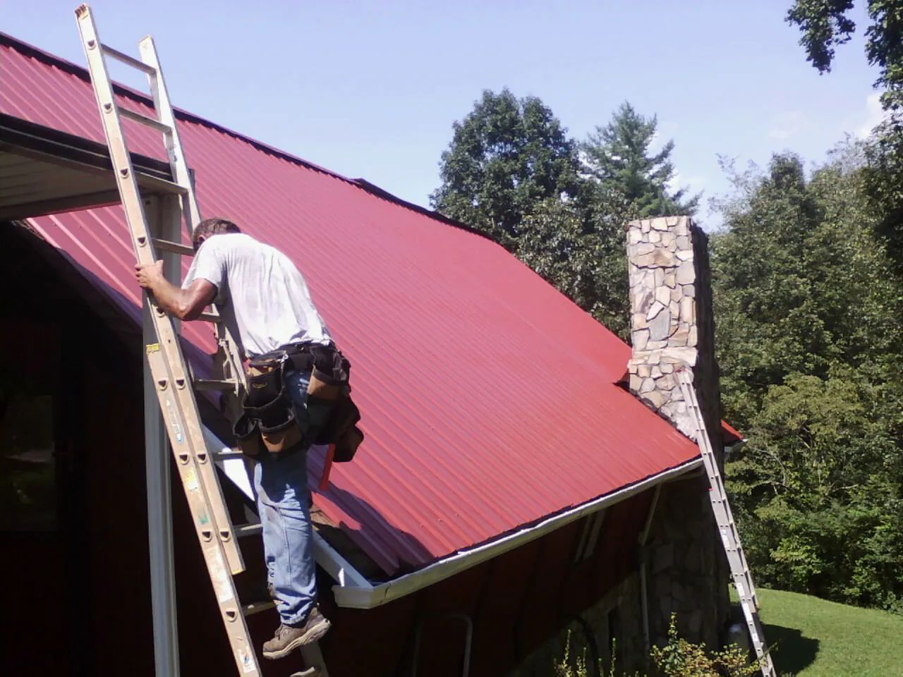 Roofer on a ladder beside a red metal roof and stone chimney — Soffit & Fascia Repair by Roofing Brothers in Wells Bridge